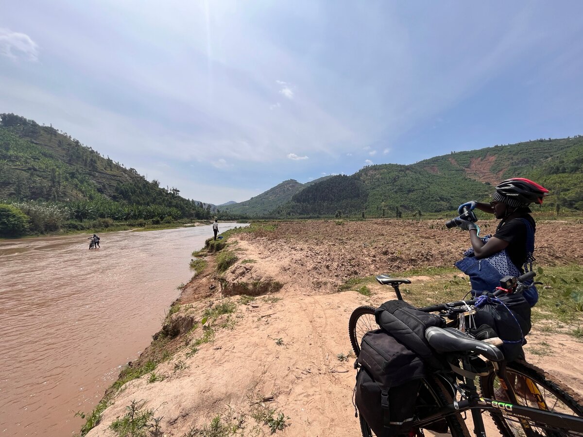 A cyclist resting beside a river with hills behind on the Congo Nile route