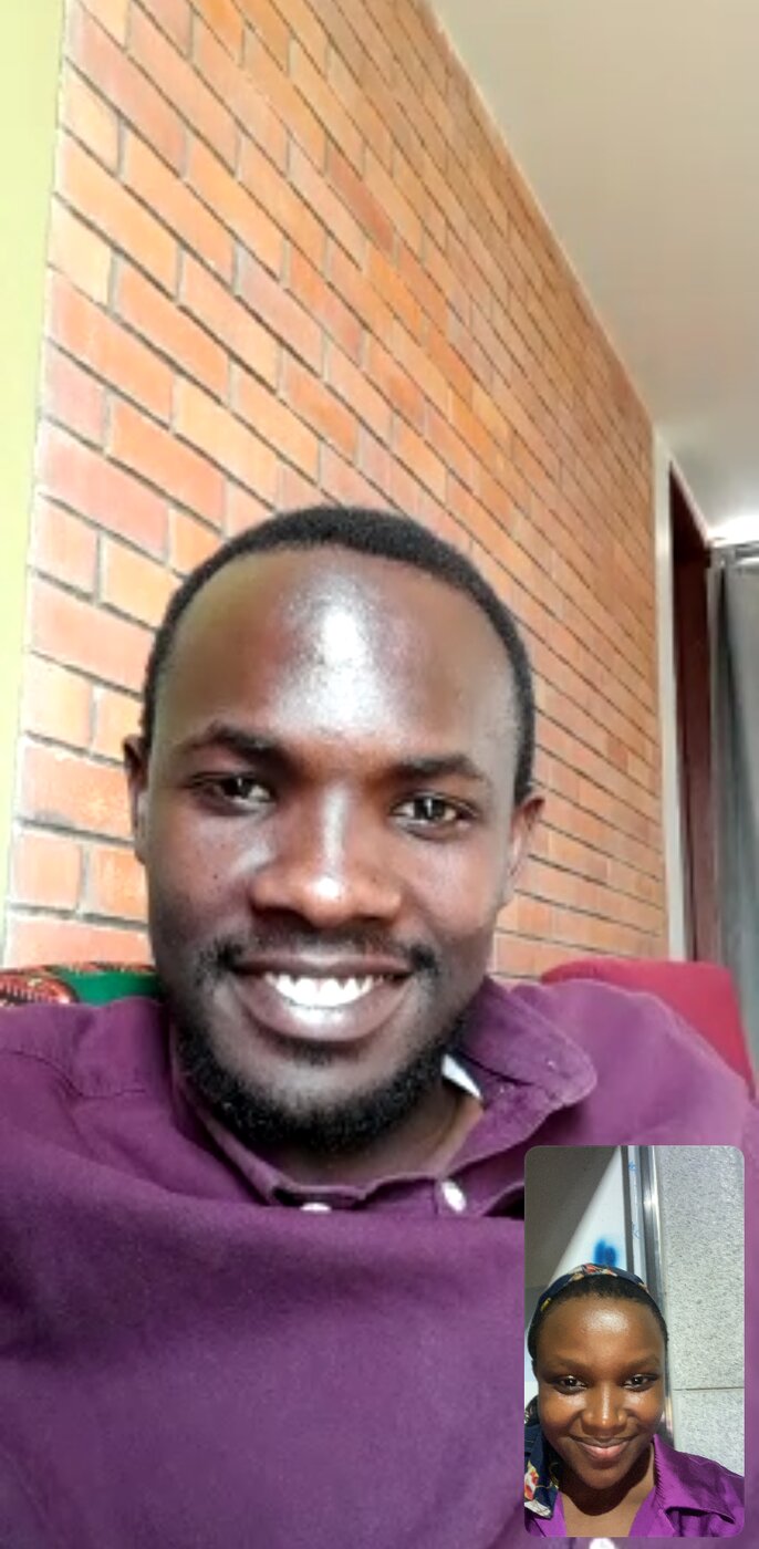 Aimable and Louise on a video call, both wearing maroon, a brick wall behind him